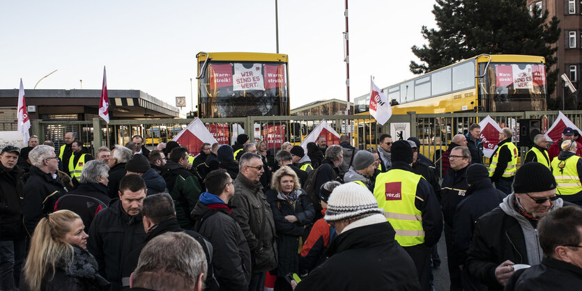 Menschen vor einer vergitterten Einfahrt, in der zwei Busse stehen