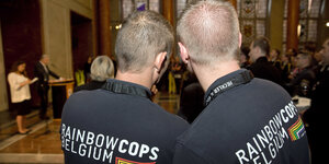 Zwei Männer sind vin hinten zu sehen, sie tragen T-Shirts mit der Aufschrift "Rainbow Cops Belgium"