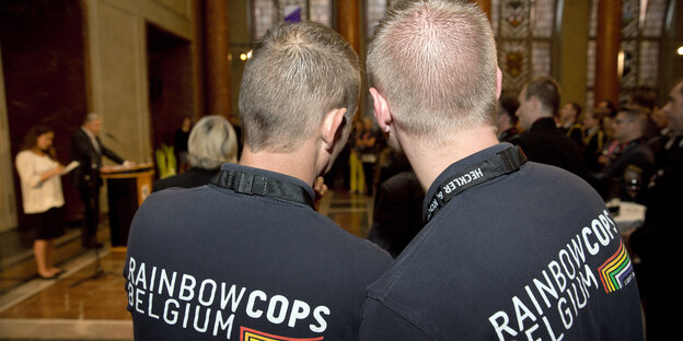 Zwei Männer sind vin hinten zu sehen, sie tragen T-Shirts mit der Aufschrift "Rainbow Cops Belgium"