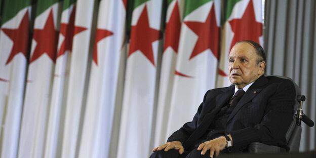Präsident Abdelaziz Bouteflika sitzt auf einem Rollstuhl. Im Hintergrund sind algerische Fahnen.