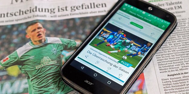 Ein Handydisplay zeigt einen Beitrag der Werder-App und liegt auf einer Ausgabe des "Weser-Kuriers".