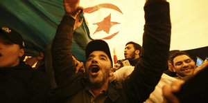 Ein Mann unter einer algerischen Flagge hebt die Arme.