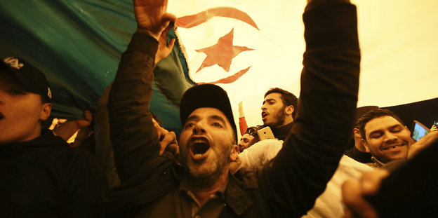 Ein Mann unter einer algerischen Flagge hebt die Arme.