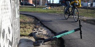 Die Lenkstange eines elektrischen Tretrollers liegt auf einem Radwege. Ein Radfahrer fährt daran vorbei