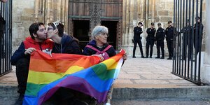 Drei Männer sitzen mit einer Regenbogenfahne vor einer Kirche im spanischen Bistum in Alcalá de Henares und protestieren gegen die Konversionstherapien.