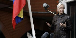 Julian Assange steht redend auf einem Balkon, links neben ihm die ecuadorianische Flagge