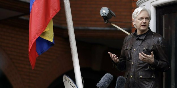 Julian Assange steht redend auf einem Balkon, links neben ihm die ecuadorianische Flagge