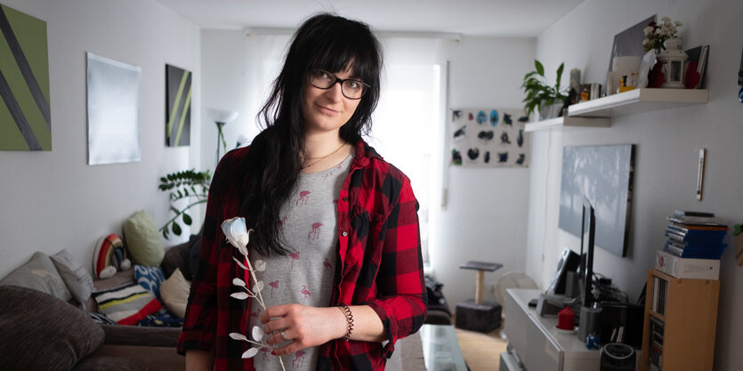 Eine Person mit langen dunklen Haaren steht in einem Zimmer und hält eine weiße Rose in der Hand