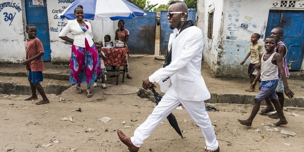 dandyhaft gekleideter Mann läuft auf einer unbefestigten Straße in Kinshasa bestaunt von Kindern und einer Frau