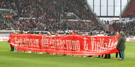 Fußballspieler tragen ein Transparent mit der Aufschrift "Gemeinsam für Erinnerung und Vielfalt" über den Rasen eines Fußballstadions.