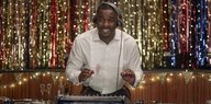 Idris Elba steht vor einem Glitzervorhang und hinter einem DJ-Pult