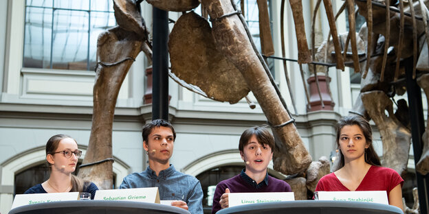 Junge Aktivist*innen sitzen vor einem Dinosaurierskelett auf einem Podium.