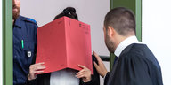 Eine Frau hält ein rotes Heft vor ihr Gesicht. Ein Mann in Anwaltsrobe berührt sie an der Schulter ubnd redet mit ihr.