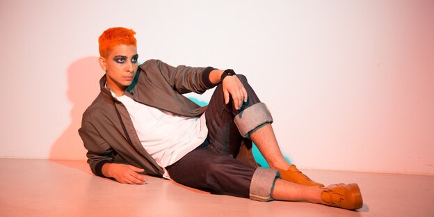Ein MAnn mit orangenen Haaren sitzt auf dem Boden und lehnt sich mit dem rechten Arm auf den Boden.
