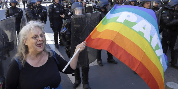 Eine FRau demonstriert mit Regenbogenflagge mit der Aufschrift „Paix“ („Frieden“), hinter ihr steht eine Polizei-Blockade