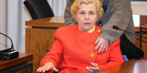 Eine Frau im Rollstuhl in roter Kleidung sitzt in einem Gerichtssaal