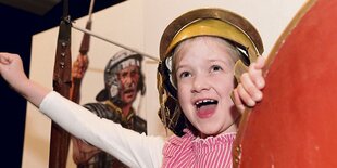 Ein Kind mit einem Legionärs-Helm steht vor der Abbildung eines Legionärs.