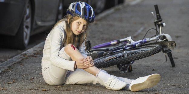 Ein Mädchen hat eine Wunde am Knie, trägt einen Fahrradhelm und sitzt vor einem Fahrrad
