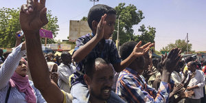 Ein Mann zeigt das Victory-Zeichen, auf seinen Schultern ein kleiner Junge. Im Hintergrund stehen viele Menschen und klatschen