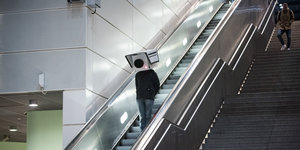 Ein teilnehmer mit einer Maske in Form einer Überwachungskamera fährt während einer Protestaktion im Bahnhof Südkreuz die Rolltreppe hoch
