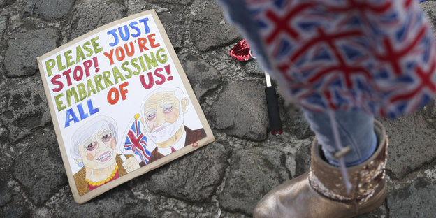 Ein Mensch steht in ein Cap mit Großbritannien-Flaggen gehüllt neben einer EU-Flagge und einem Plakat mit einer Zeichnung von Theresa May und Jeremy Corbyn und der Aufschrift: „Bitte hört auf, uns zu blamieren“
