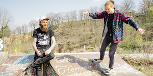 Tobias Burdukats sitzt auf einer Halfpipe, ein junger Mann fährt dort mit seinem Skateboard