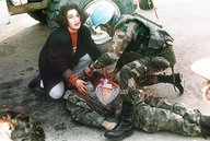 Ein Mann in Militärkleidung liegt am Boden, er ist verletzt, eine Frau und ein Mann knien neben ihm