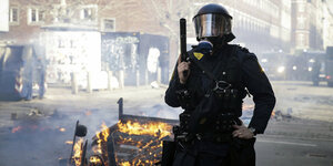Ein Polizist in Riotuniform mit einer gezogenen Waffe vor einer kleinen brennenden Barrikade