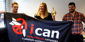 Eine Frau und zwei Männer halten ein Transparent in die von Ican in die Höhe. Die Frau hält in ihrer linken Hand zudem eine Flascheauch