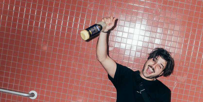 Ein junger Mann mit Schnurrbart und irrem Blick hält eine Flasche Eierlikör in der Hand