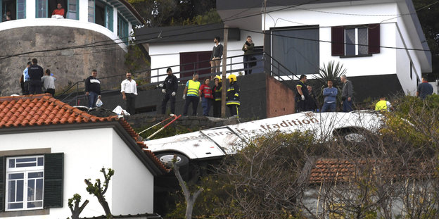 Ein weißer Bus liegt auf dem Dach eines Hauses am Hang, oberhalb stehen Menschen zum Teil in Warnwesten und blicken auf den Bus hinab.
