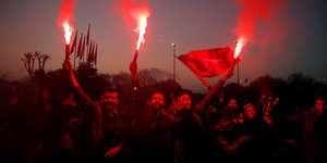 Eine Gruppe von Menschen halten feiernd rote Fahnen und Pyrotechnik hoch, im Hintergrund der Horizont bei Dämmerung.