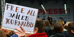 eine Frau hält ein A4-Blatt hoch, darauf steht: „free all jailed journalists“