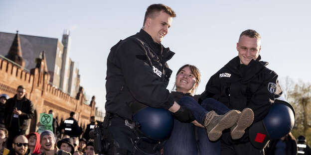 Zwei Polizisten tragen eine Klimaaktivist*in weg. Alle lächeln