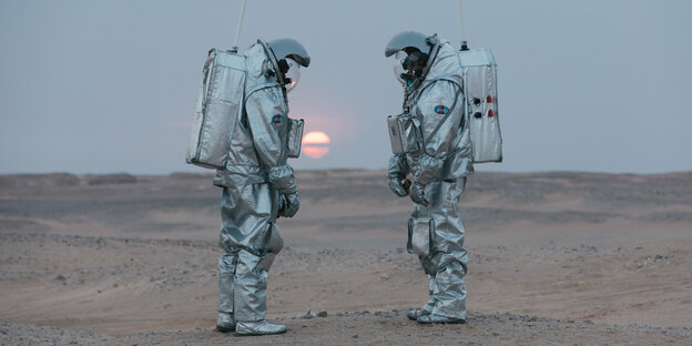Zwei Menschen in Astronaut-Anzügen in der Wüste bei untergehender Sonne.