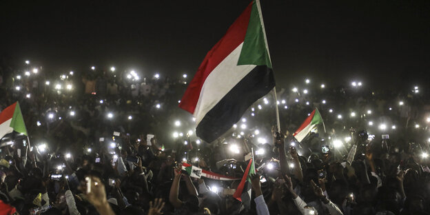 Menschen halten bei Nacht Smartphones in die Luft, in der Mitte des Bildes hält jemand die sudanesische Fahne hoch
