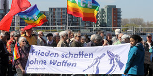 Mehrere Menschen demonstrieren in Rostock für den Frieden bei einem Ostermarsch. Sie haben eine bunte Regenbogenfahne mit der Aufschrift "Pace" in der Hand.