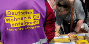 Ein mensch trägt eine Weste mit der Aufschrift „Deutsche Wohnen und Co. enteignen! Hier unterschreiben“, eine Frau stützt sich davor auf einen Tisch und unterschreibt