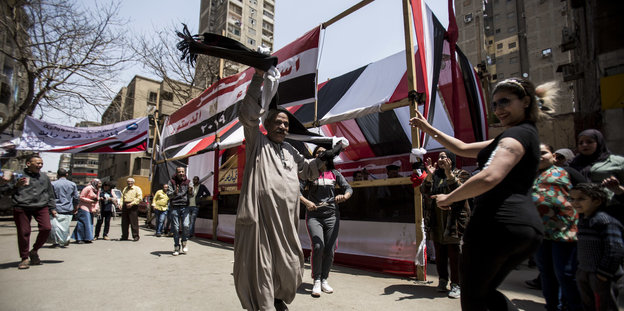 Eine Straße in Kairo mit ägyptischen Flaggen, im Vordergrund ein älterer Mann und eine junge Frau, die tanzen