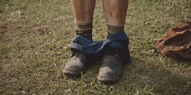 Männliche Beine in Arbeitsschuhen, auf denen eine heruntergelassene Unterhose liegt, daneben im Gras liegt ein T-Shirt.