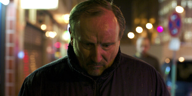 Schauspieler Rainer Bock als Scholl, im Gegenlicht auf der Straße. Sein Gesicht in Nahaufnahme und im Dunkeln, hinter ihm die blinkenden Straßenlichter, nachts.