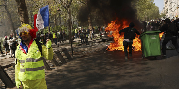 Ein Gelbwesten-Demonstrant, dahinter zerrt jemand eine Mülltonne ins Feuer