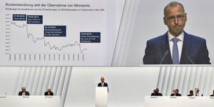 Ein Mann, Werner Baumann, spricht auf einem weißen Podium vor einer großen Leinwand, auf der wiederum er zu sehen ist