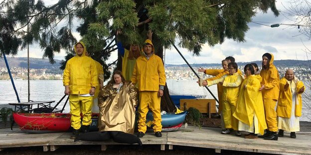Viele Schauspieler in gelben Regenjacken am Ufer eines Sees.