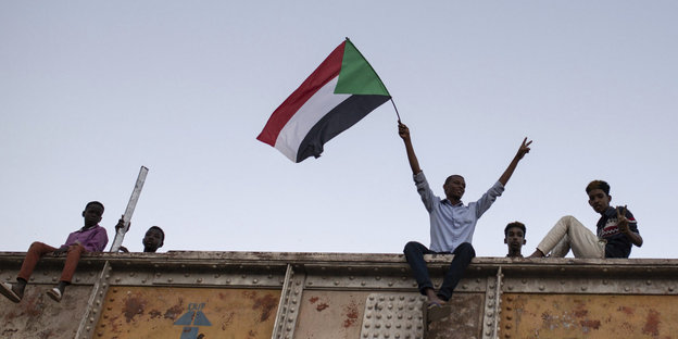 Ein Mann sitzt auf einem Container und hält eine sudanesische Flagge hoch