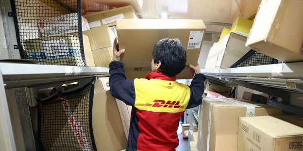 Eine DHL-Mitarbeiterin belädt ein Lieferfahrzeug mit Paketen. Sie ist von hinten zu sehen, wie sie ein Paket in einen Lieferraum mit vielen Paketen hebt.