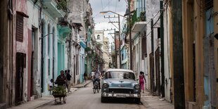 Eine Straßenszene in Havanna zeigt ein geparktes Auto, mehrere Fußgänger und bunte Häuser links und rechts