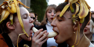 Zwei Personen haben Bananenschalen auf dem Kopf und beißen von zwei Seiten in eine geschälte Banane.