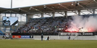 Auf einer Tribüne zünden Hooligans Bengalos, an der Bande steht "Ruhe in Frieden, Tommy"