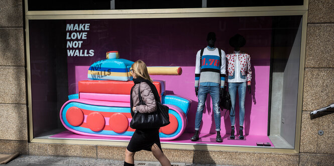 eine Frau geht an einem Schaufenster vorbei, darin ein pinker Panzer und der Textzug: Make Love not walls